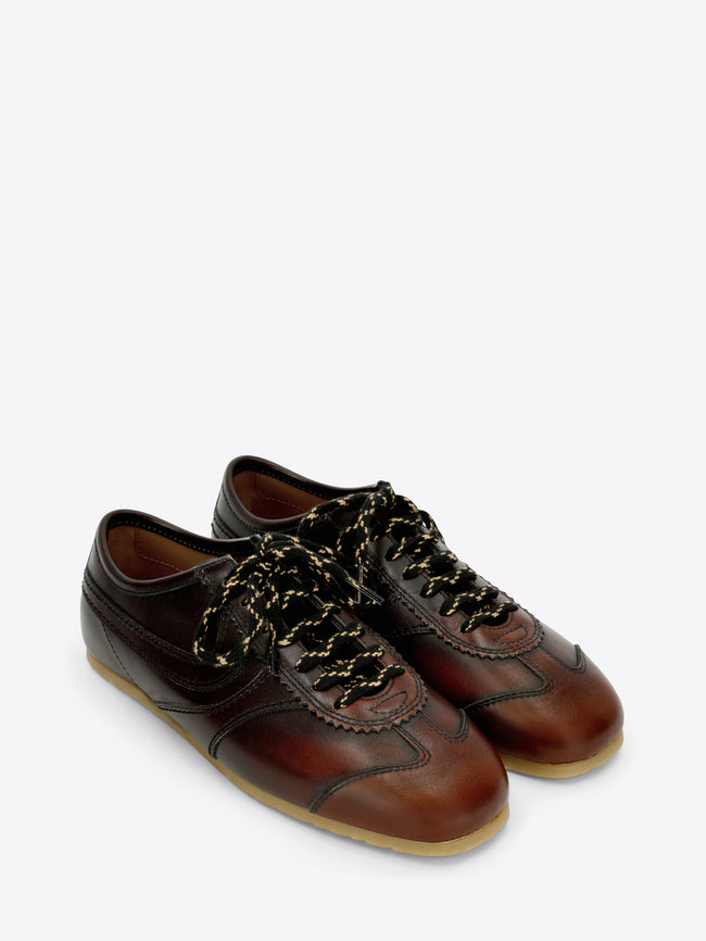 A pair of dark brown leather low-top sneakers with decorative laces, a brown leather toe cap, and a light tan rubber sole, shown against a light gray background.