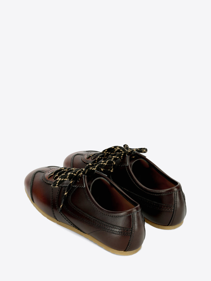 A pair of dark brown leather low-top sneakers with decorative laces, a brown leather toe cap, and a light tan rubber sole, shown against a light gray background.