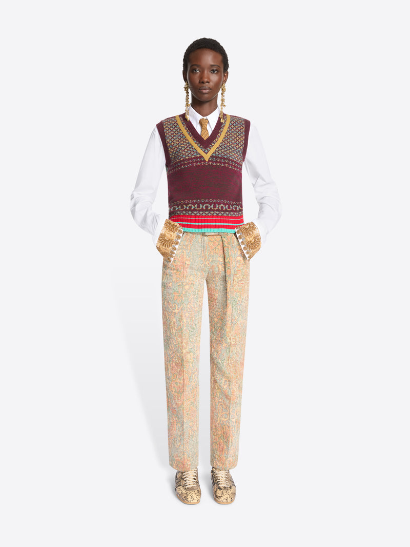A model stands against a light gray background wearing a white long-sleeve shirt, a burgundy patterned knit sweater vest, a tie, beige patterned trousers, and gold snakeskin shoes.