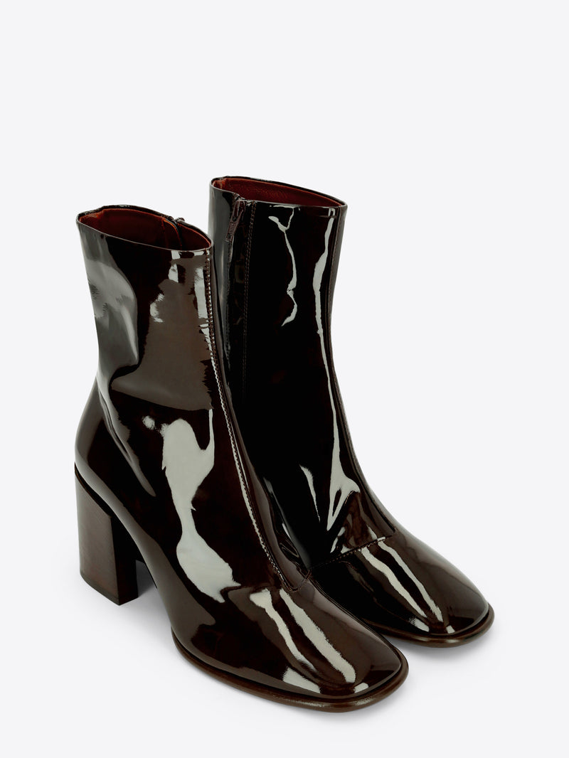 A pair of dark olive patent leather ankle boots with a rounded square toe and chunky block heel on a light gray background.
