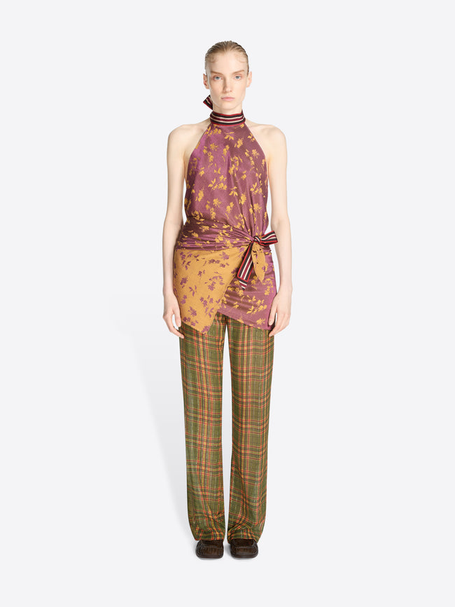 A model stands against a light gray background wearing a sleeveless halter top with a purple and orange abstract print, tied at the waist, paired with green and orange plaid trousers.