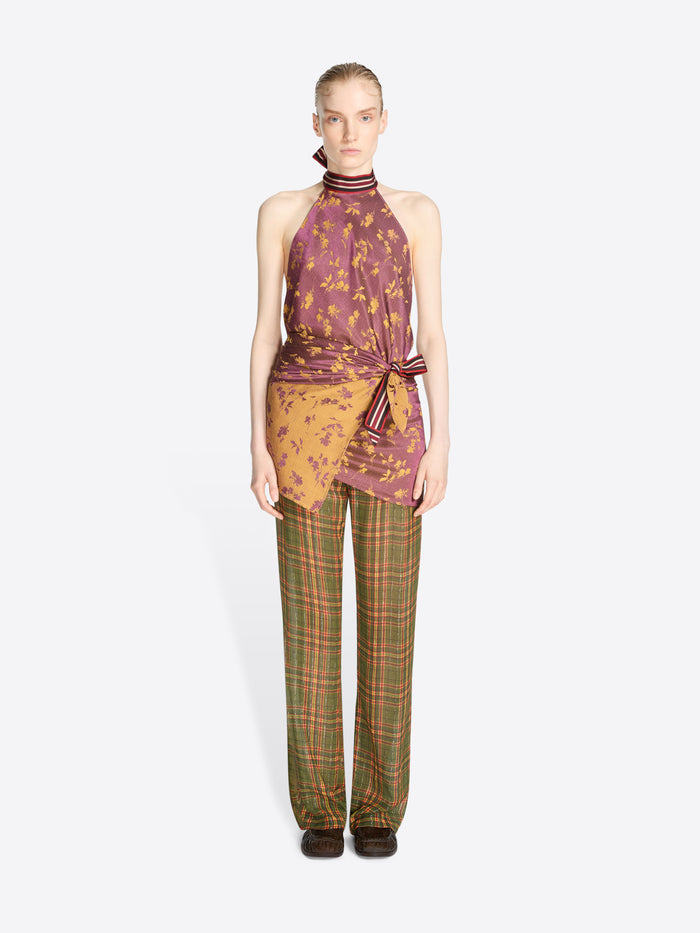 A model stands against a light gray background wearing a sleeveless halter top with a purple and orange abstract print, tied at the waist, paired with green and orange plaid trousers.