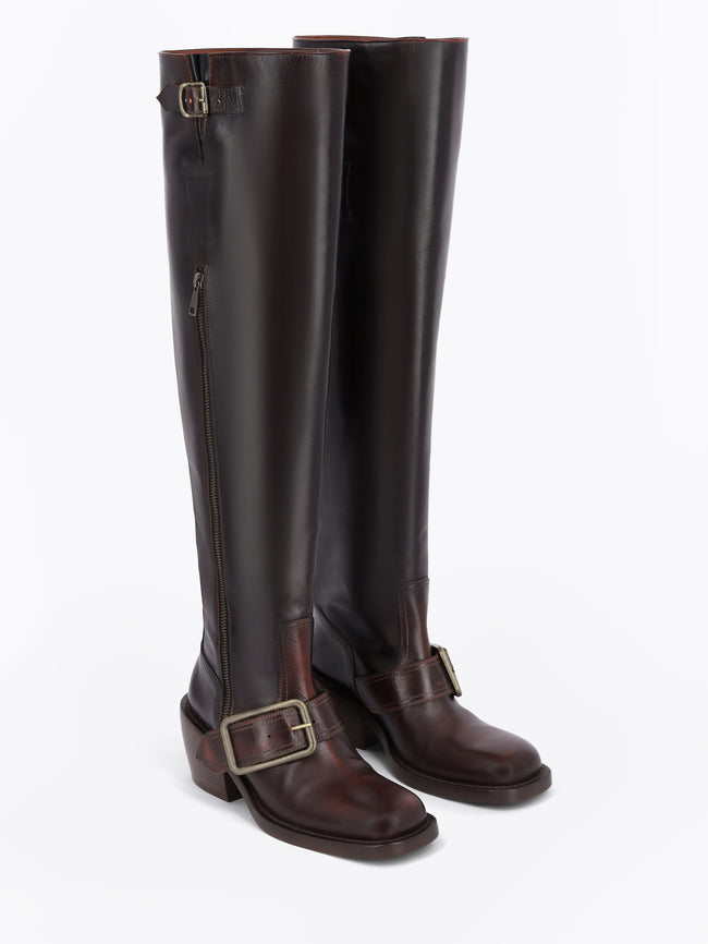A pair of dark brown leather knee-high boots with a side zipper, a decorative strap with a gold buckle near the top, and a matching buckle on the lower shaft, shown against a light gray background.