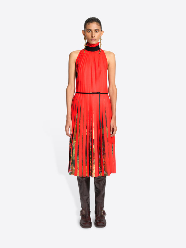 A model stands against a light gray background wearing a sleeveless high-neck red dress with a pleated skirt and a thin black belt, paired with dark brown knee-high boots.