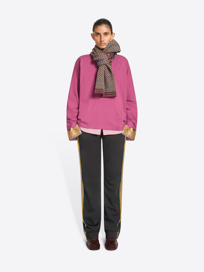 A model stands against a light gray background wearing a bright pink sweatshirt, pink shirt with gold patterned cuffs, a patterned scarf, black track pants with side stripes, and dark brown shoes.