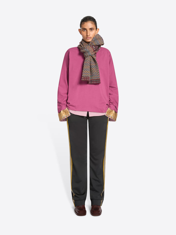 A model stands against a light gray background wearing a bright pink sweatshirt, pink shirt with gold patterned cuffs, a patterned scarf, black track pants with side stripes, and dark brown shoes.