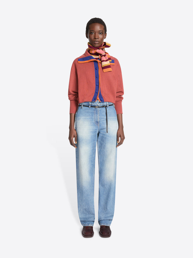 A model stands against a light gray background wearing a coral-orange cropped cardigan with a blue and orange striped collar detail, a multicolored striped neck scarf, light-wash blue jeans, and dark brown shoes.