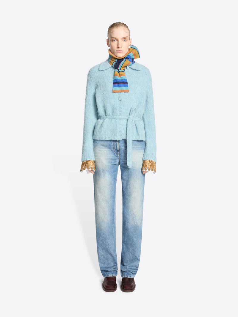 A model stands against a light gray background wearing a light blue belted jacket, white shirt  with gold lace cuff accents, light-wash blue jeans, and a multicolored striped neck scarf.