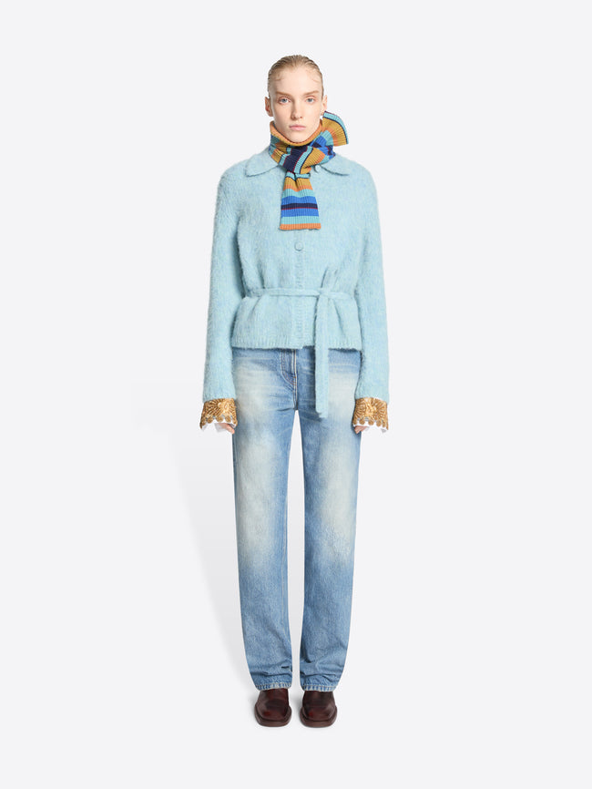 A model stands against a light gray background wearing a light blue belted jacket, white shirt  with gold lace cuff accents, light-wash blue jeans, and a multicolored striped neck scarf.