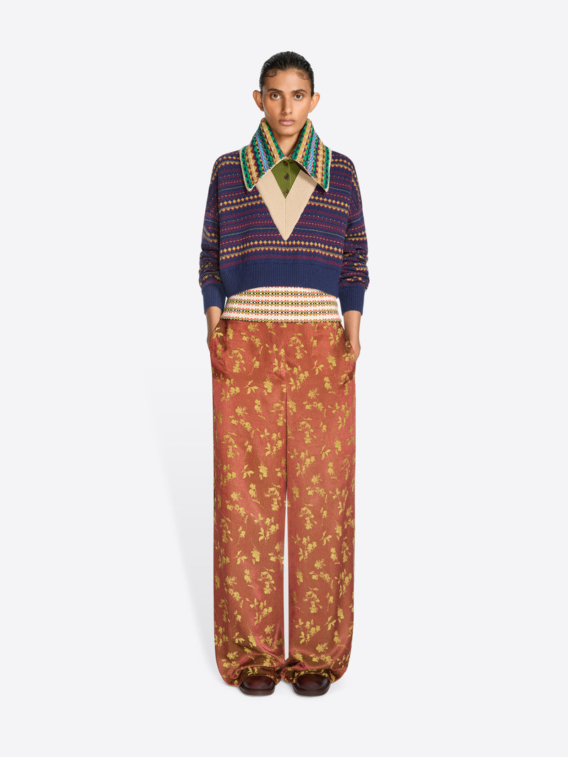 A model stands against a light gray background wearing a cropped knit sweater with a multicolor patterned collar, paired with wide-leg rust-colored pants featuring a gold floral print.