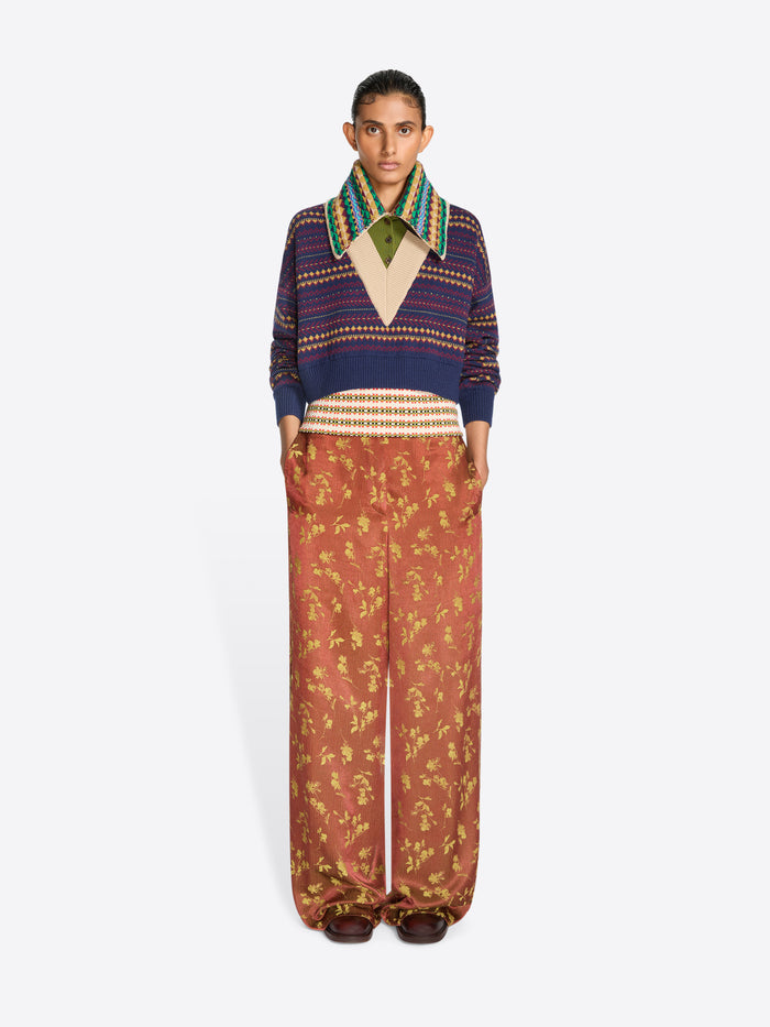 A model stands against a light gray background wearing a cropped knit sweater with a multicolor patterned collar, paired with wide-leg rust-colored pants featuring a gold floral print.