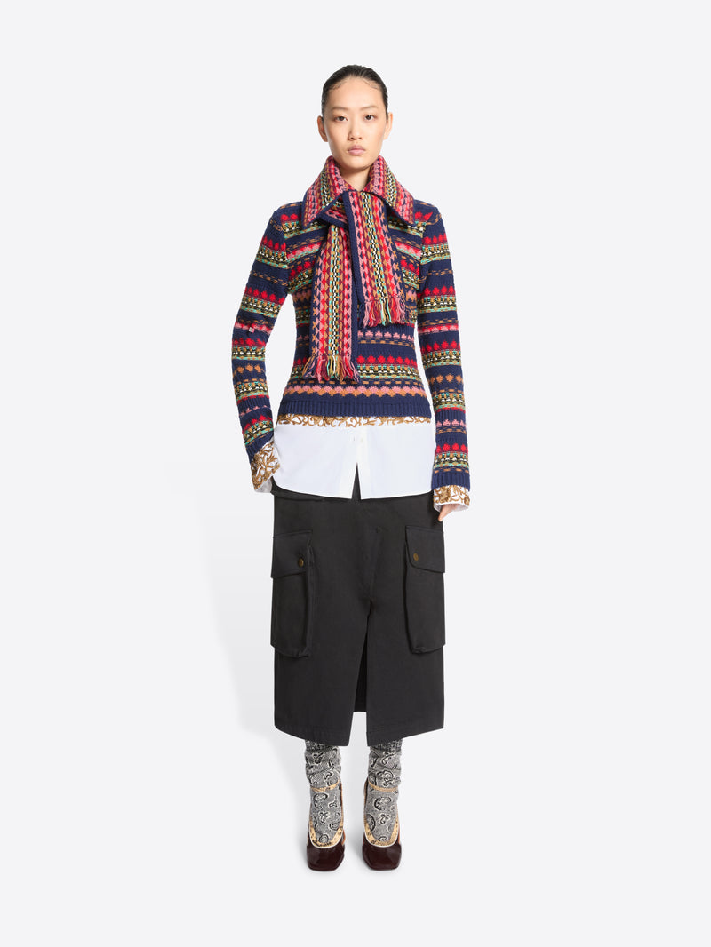 A model stands against a light gray background wearing a colorful patterned knit sweater, a knit collar with a colorful geometric pattern, a white shirt tucked in, black cargo-style skirt, patterned socks, and dark heels.