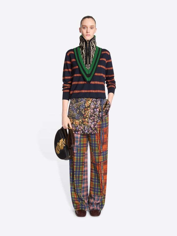 A model stands against a light gray studio background wearing a dark striped sweater, a multicolored knitted neck warmer, wide-leg plaid trousers, carrying a small black bag with gold embroidery.