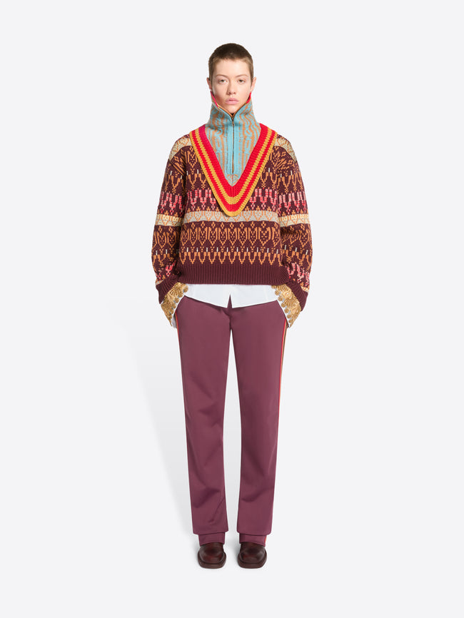 A model stands against a light gray studio background wearing a patterned knit sweater, a multicolored knitted neck warmer, burgundy pants, and dark brown shoes.