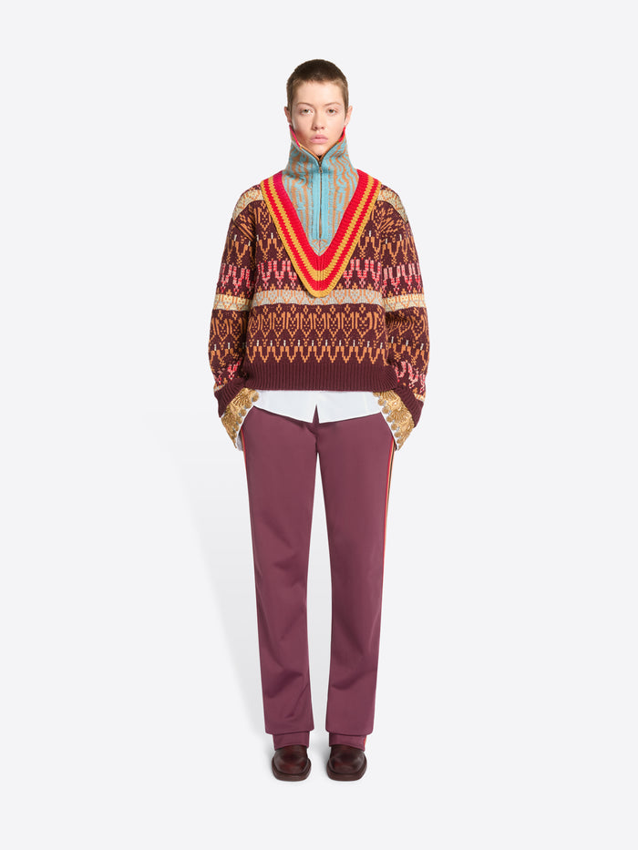 A model stands against a light gray studio background wearing a patterned knit sweater, a multicolored knitted neck warmer, burgundy pants, and dark brown shoes.