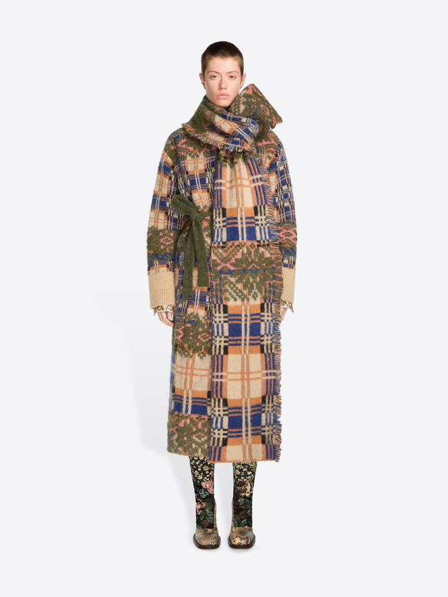 A model stands against a light gray background wearing a long, wide patterned coat with a matching scarf and patterned boots. 