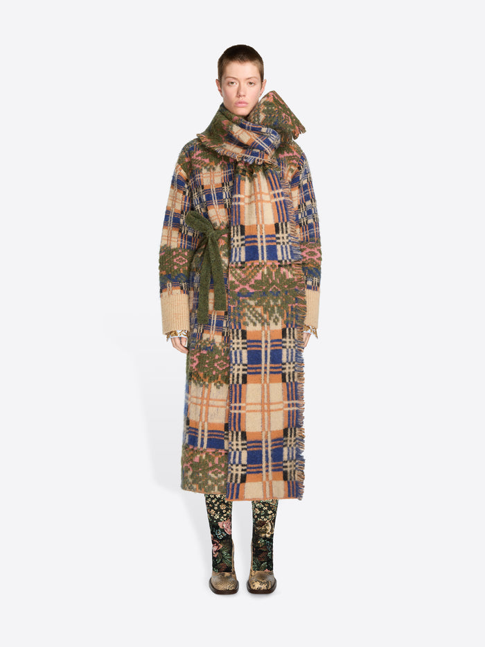 A model stands against a light gray background wearing a long, wide patterned coat with a matching scarf and patterned boots. 
