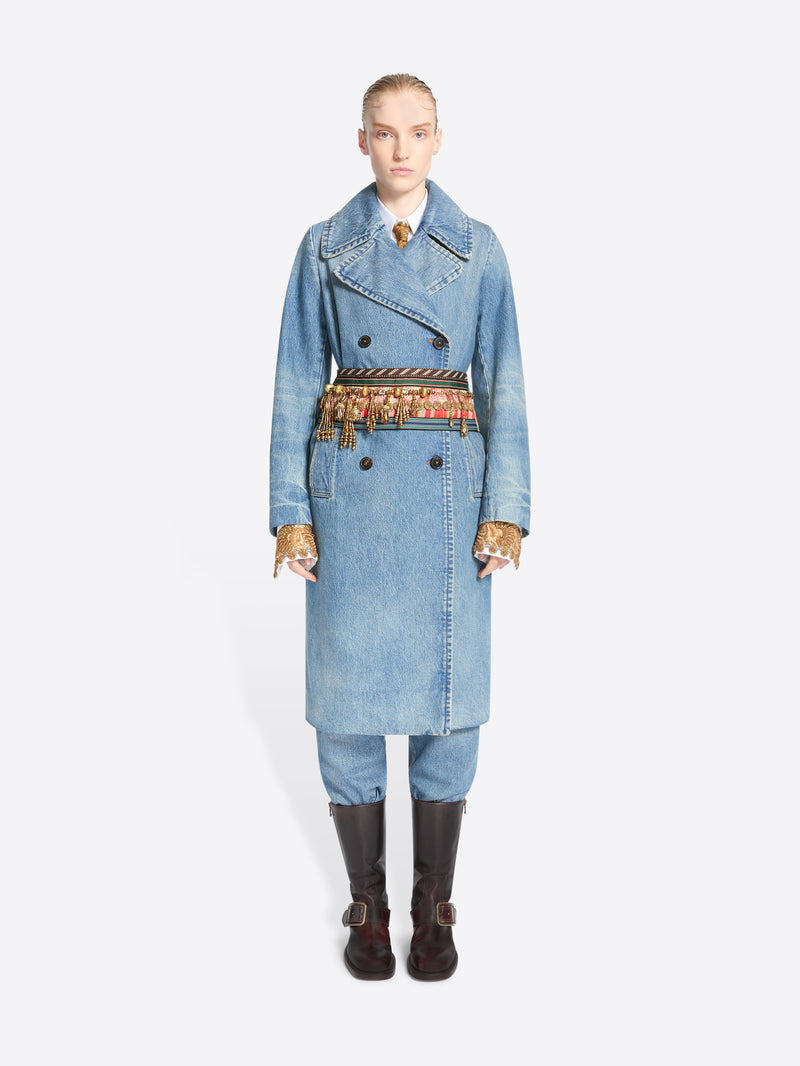 Full-length model wearing a light blue denim trench coat and matching trousers, with an old brass charm necklace styled as a belt and dark brown knee-high boots.