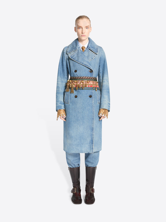 Full-length model wearing a light blue denim trench coat and matching trousers, with an old brass charm necklace styled as a belt and dark brown knee-high boots.