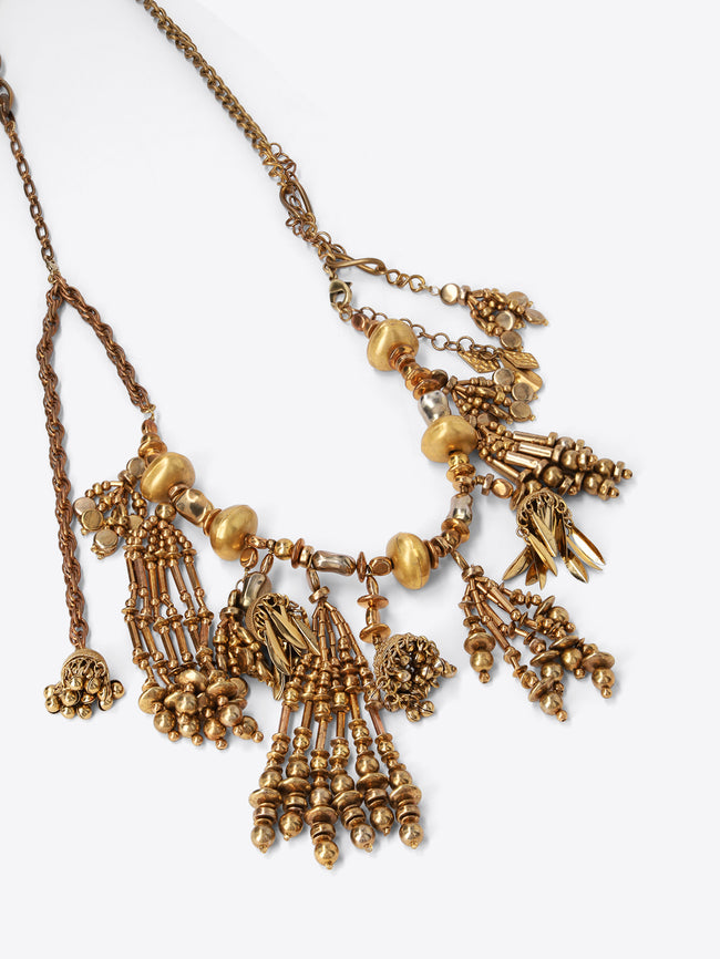 Close-up of an old brass necklace with rounded beads and cascading tassel, leaf, and bead charms on a light gray background.