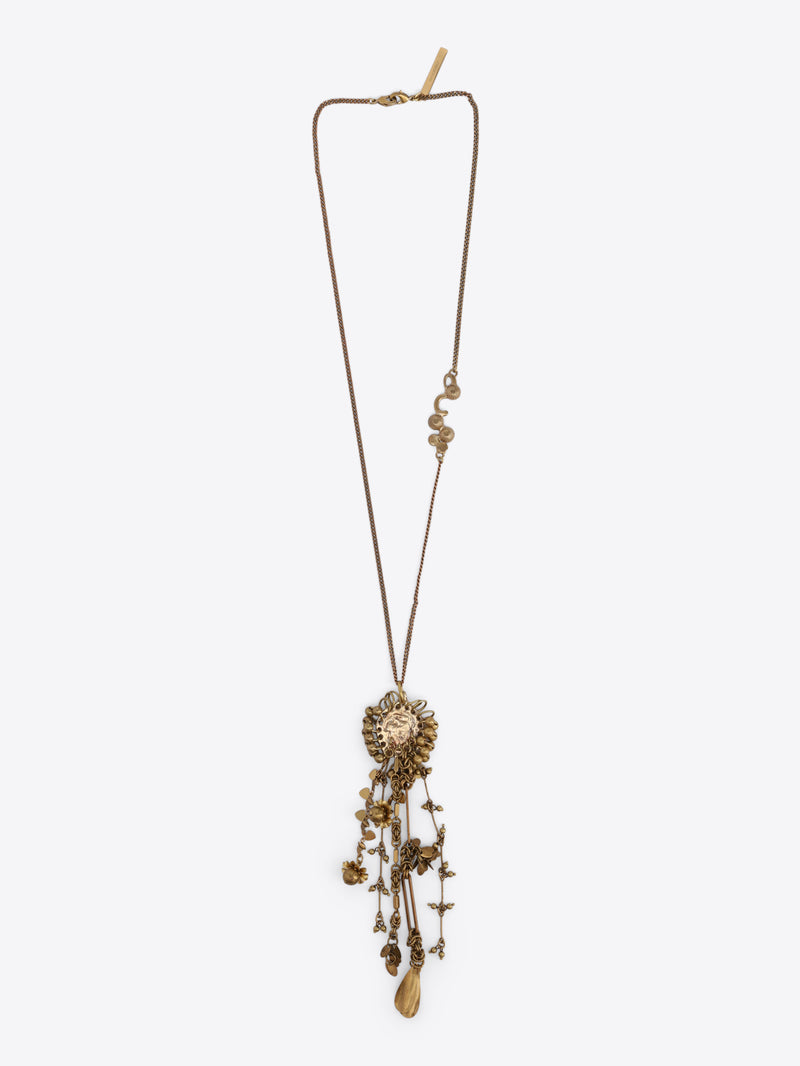 Long old brass necklace with an embossed medallion pendant and cascading floral, bead, and teardrop charms on a light gray background.