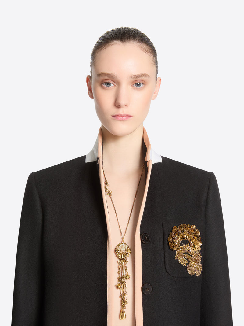 Front-facing model wearing a long old brass necklace with an ornate medallion and cascading charms, paired with a black blazer featuring gold embroidery.