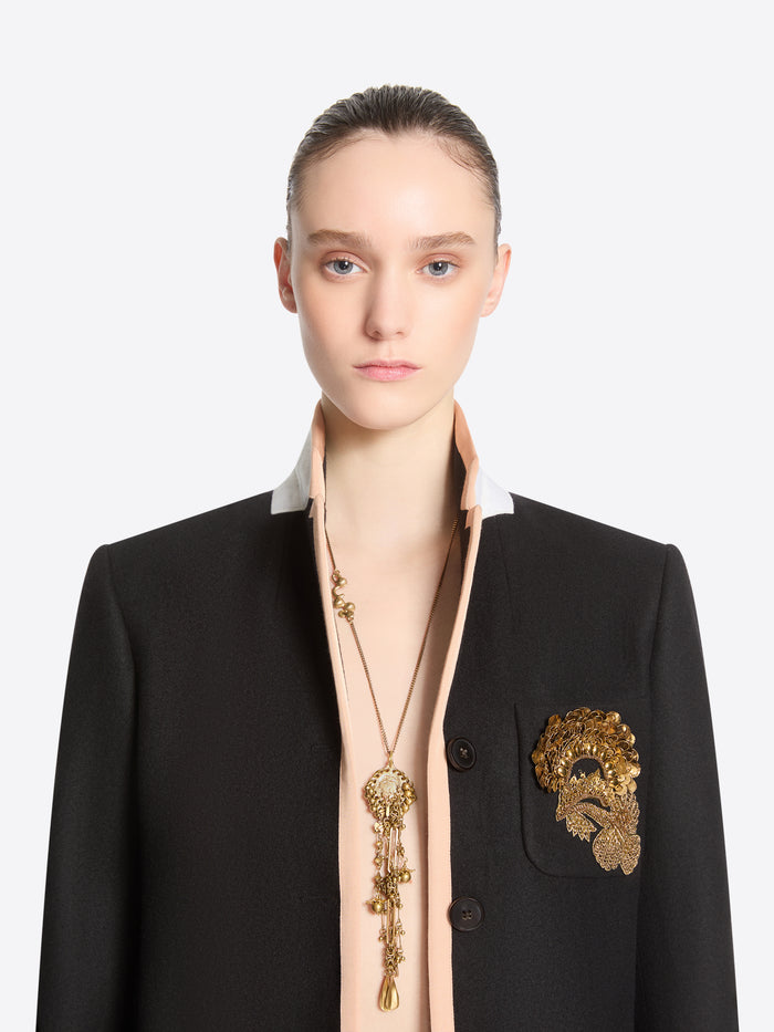 Front-facing model wearing a long old brass necklace with an ornate medallion and cascading charms, paired with a black blazer featuring gold embroidery.