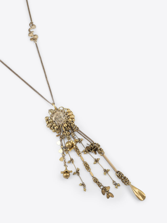 Close-up of an old brass necklace with an embossed medallion and cascading floral, heart, bead, and teardrop charms on a light gray background.
