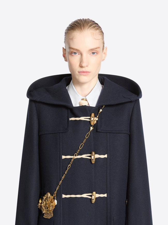 Front-facing model wearing a navy duffle coat with a long old brass necklace draped diagonally and finished with an ornate charm cluster.
