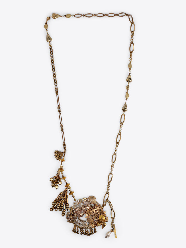 Long old brass necklace with mixed chain links and an ornate cluster of floral, medallion, bead, and tassel charms on a light gray background.
