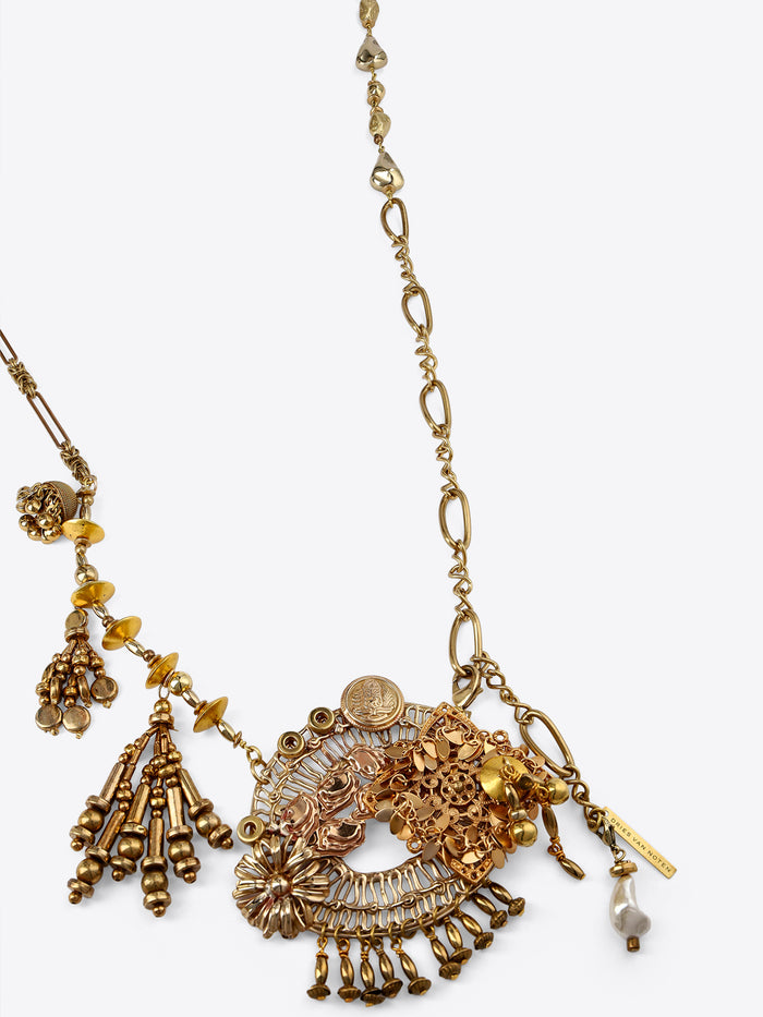 Close-up of an old brass necklace with mixed links, an ornate floral and medallion charm cluster, beaded tassels, and a pearl drop on a light gray background.