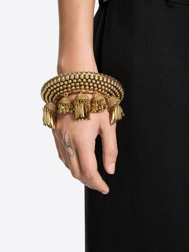 Close-up of a hand wearing an ornate old brass cuff bracelet with a beaded surface and dangling leaf and bead charms against a black garment.