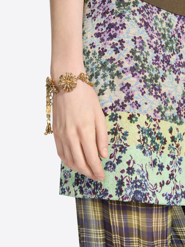 Close-up of a hand wearing an old brass charm bracelet with sculpted flowers and dangling leaf and bud charms against a multicolored floral garment.