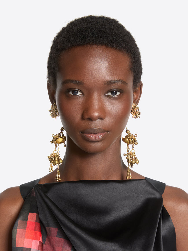 A close-up portrait of a model wearing large, ornate gold dangle earrings with floral and leaf motifs, paired with a black satin top featuring a red and black plaid pattern on the shoulder.