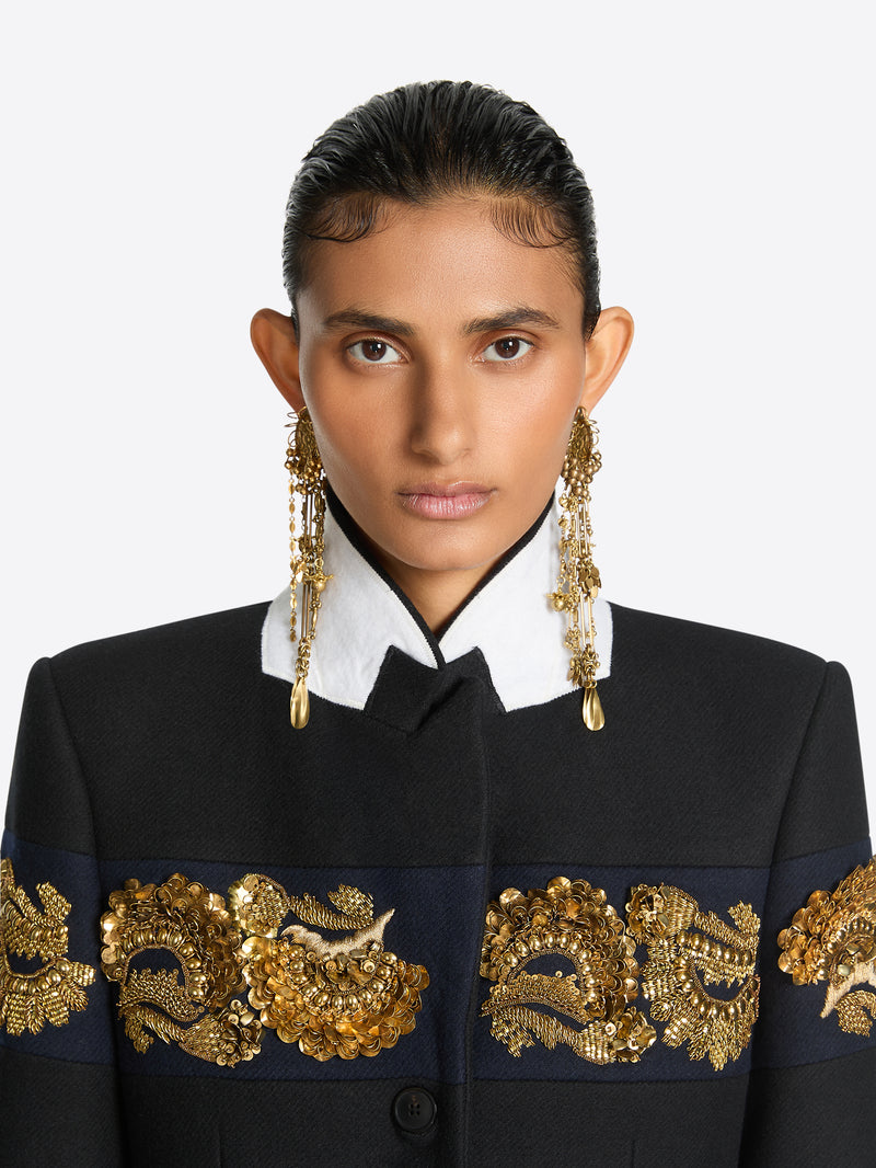 Front-facing model with slicked-back hair wearing long old brass drop earrings with cascading chains and teardrop pendants, paired with a black jacket featuring a white collar and ornate gold embroidery.