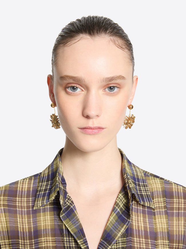 Front-facing model with slicked-back hair wearing gold-toned flower drop earrings and a multicolored plaid shirt.