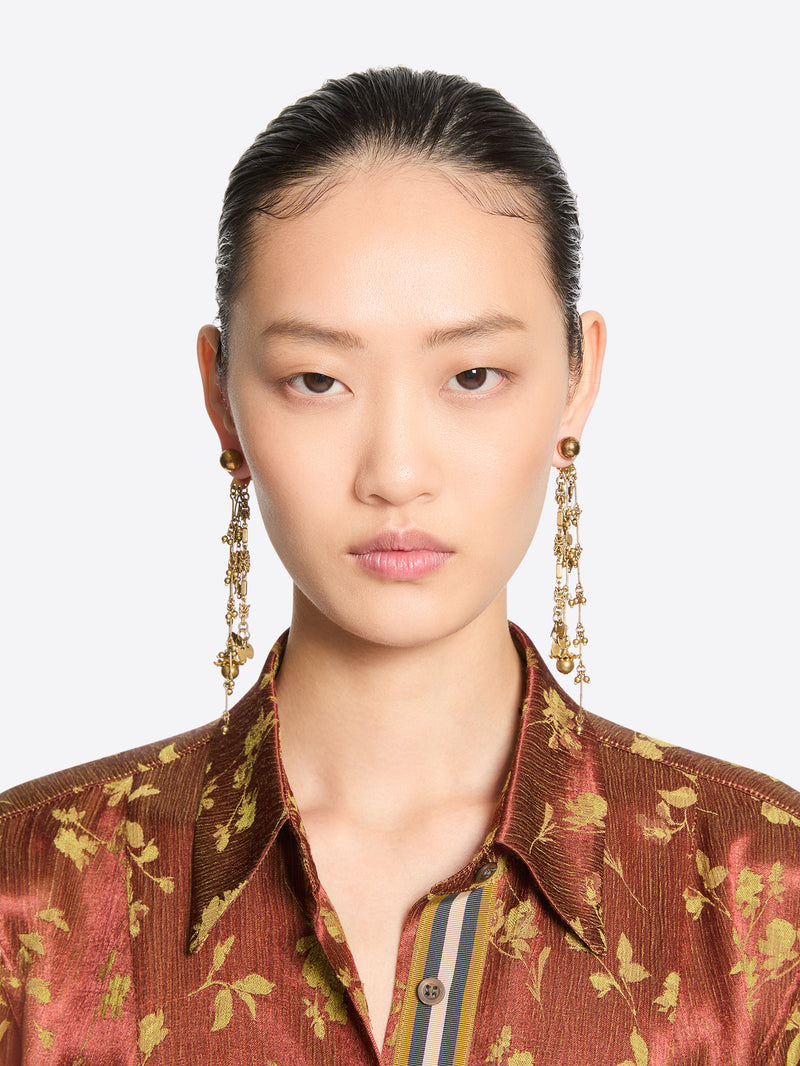 Front-facing model with slicked-back hair wearing long old brass drop earrings with cascading chains and charms, paired with a rust-toned floral shirt.