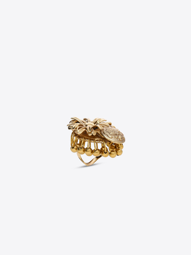Gold-toned flower ring with layered petals and a dangling embossed medallion charm on a light gray background.