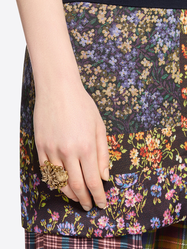 Close-up of a hand wearing a gold-toned flower ring with a dangling embossed medallion against multicolored floral-patterned fabric.