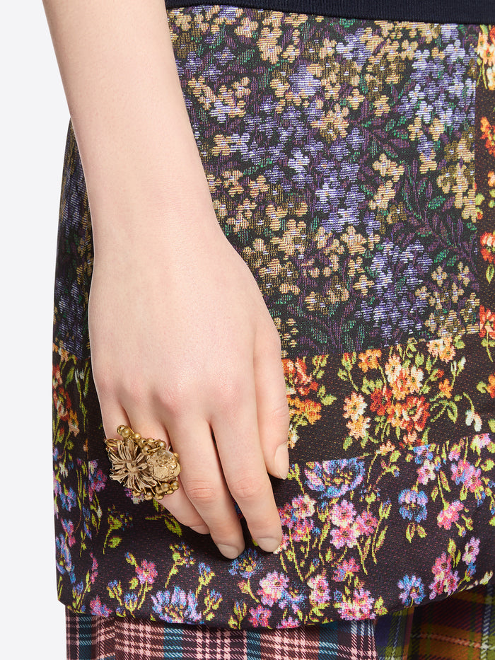 Close-up of a hand wearing a gold-toned flower ring with a dangling embossed medallion against multicolored floral-patterned fabric.