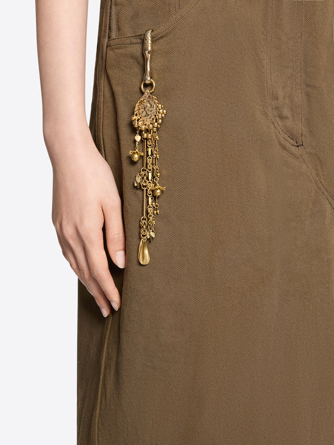 Close-up of an old brass decorative clip with cascading chains and charms attached to an olive-brown garment beside the model’s hand.
