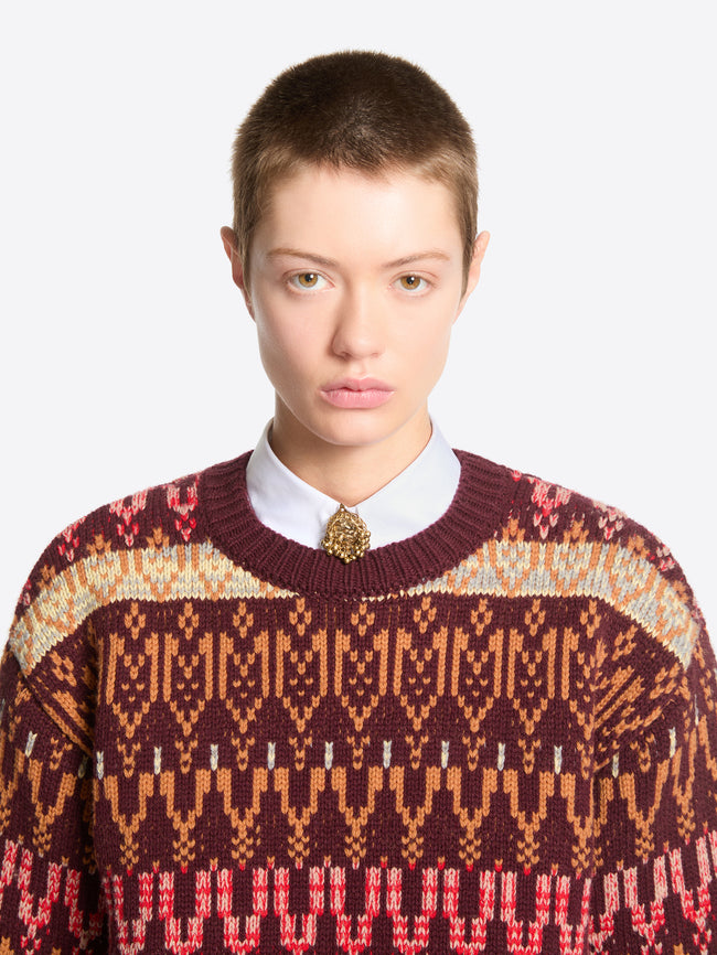 Front-facing model with short hair wearing a burgundy patterned knit sweater over a white collared shirt, accented with a gold-toned grape-cluster button cover.