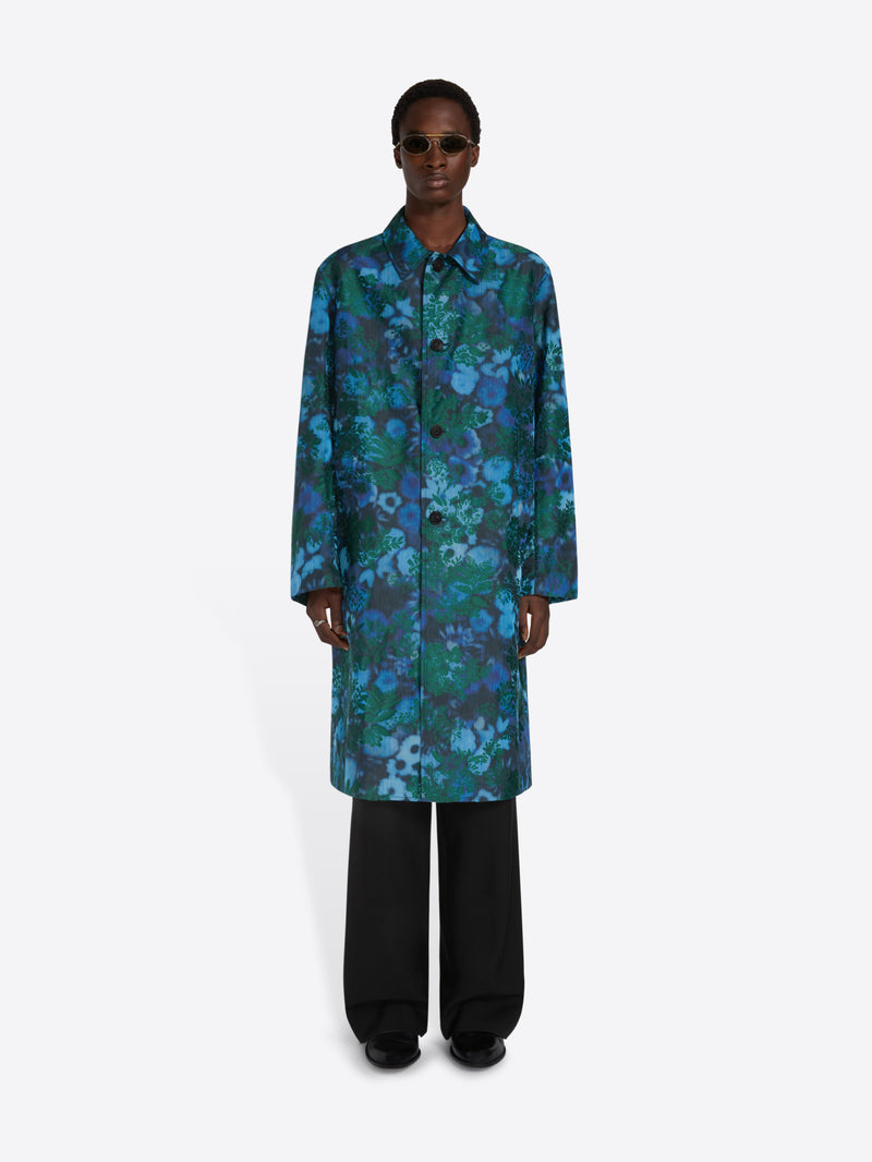 A model wearing a long-sleeved, knee-length button-up coat with an abstract blue, green, and teal floral pattern, paired with black wide-leg trousers and sunglasses, set against a light gray background.
