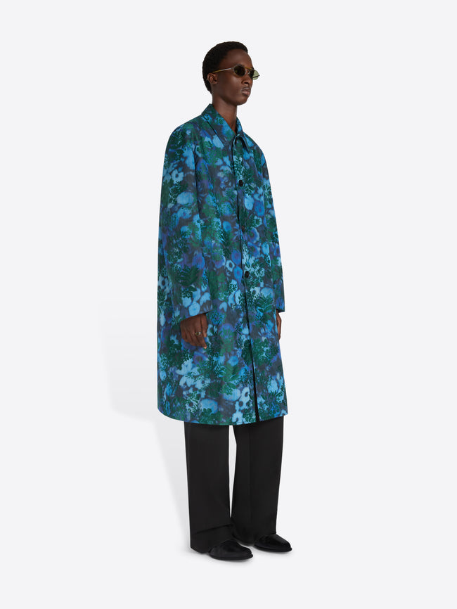 A model wearing a long-sleeved button-up shirt jacket with a vibrant blue and green floral pattern, paired with black trousers and sunglasses, standing against a gray background.