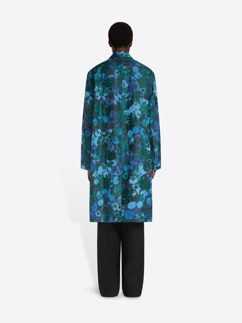 A model shown from the back wearing a long-sleeved, knee-length coat with a vibrant blue and green floral pattern, paired with black pants, against a plain light gray background.