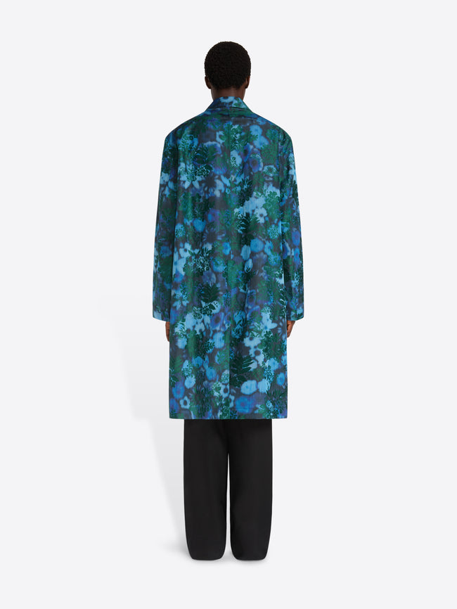 A model shown from the back wearing a long-sleeved, knee-length coat with a vibrant blue and green floral pattern, paired with black pants, against a plain light gray background.