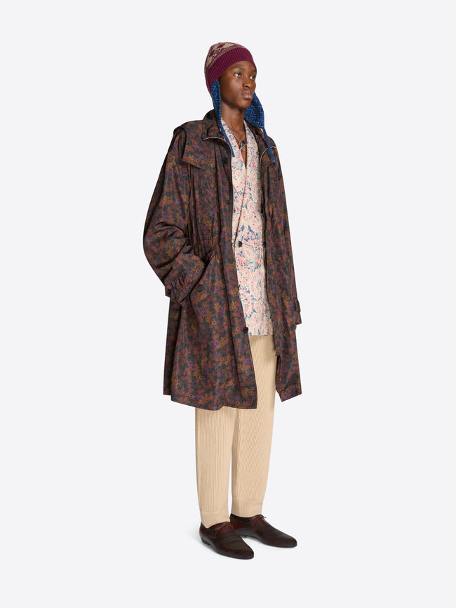 Person wearing a long patterned floral coat, floral shirt, hat and beige pants on a white background