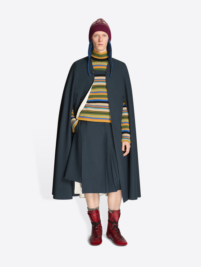 Person wearing a dark cape, striped sweater, skirt, hat and red boots on a white background