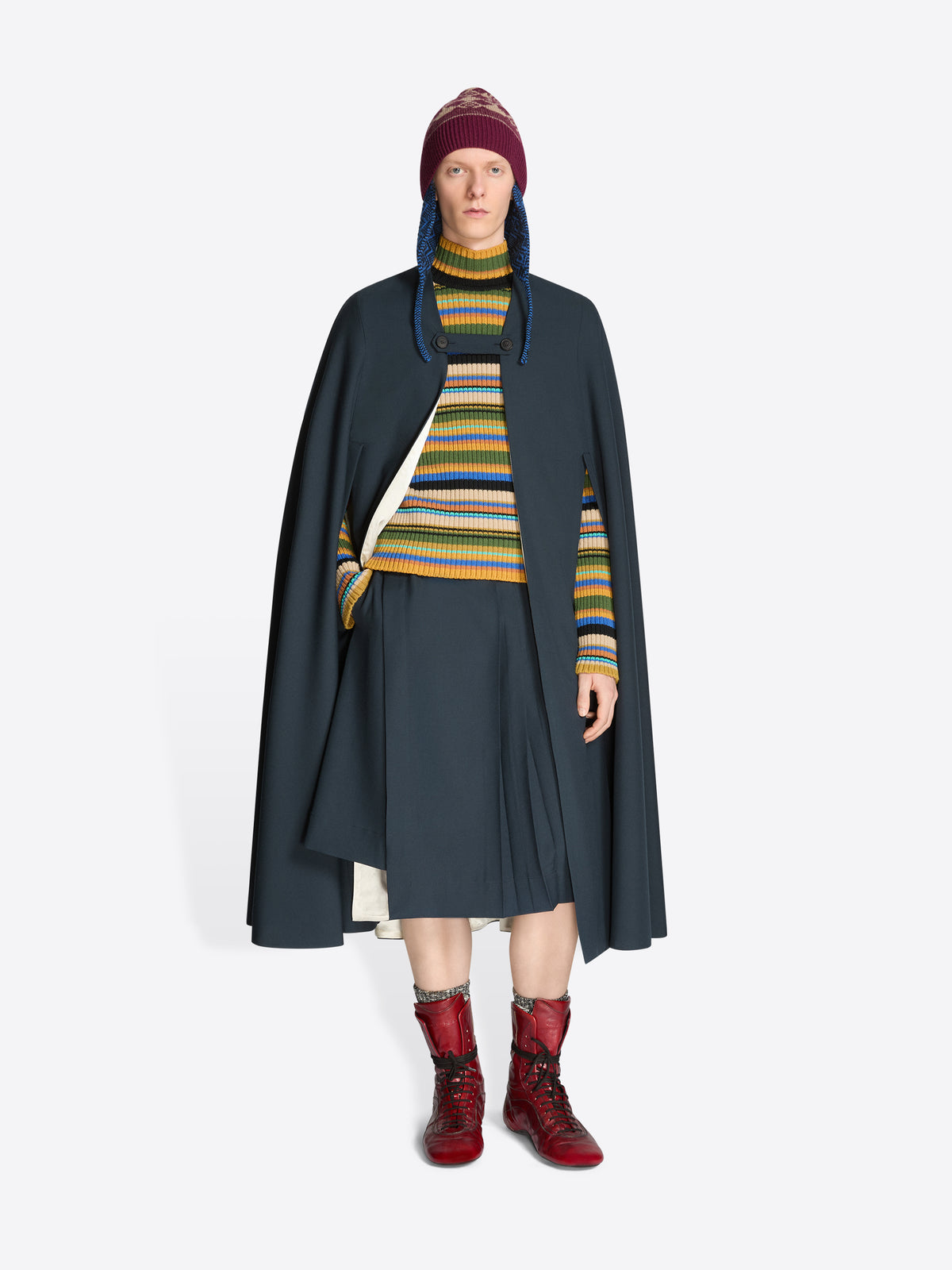 Person wearing a dark cape, striped sweater, skirt, hat and red boots on a white background