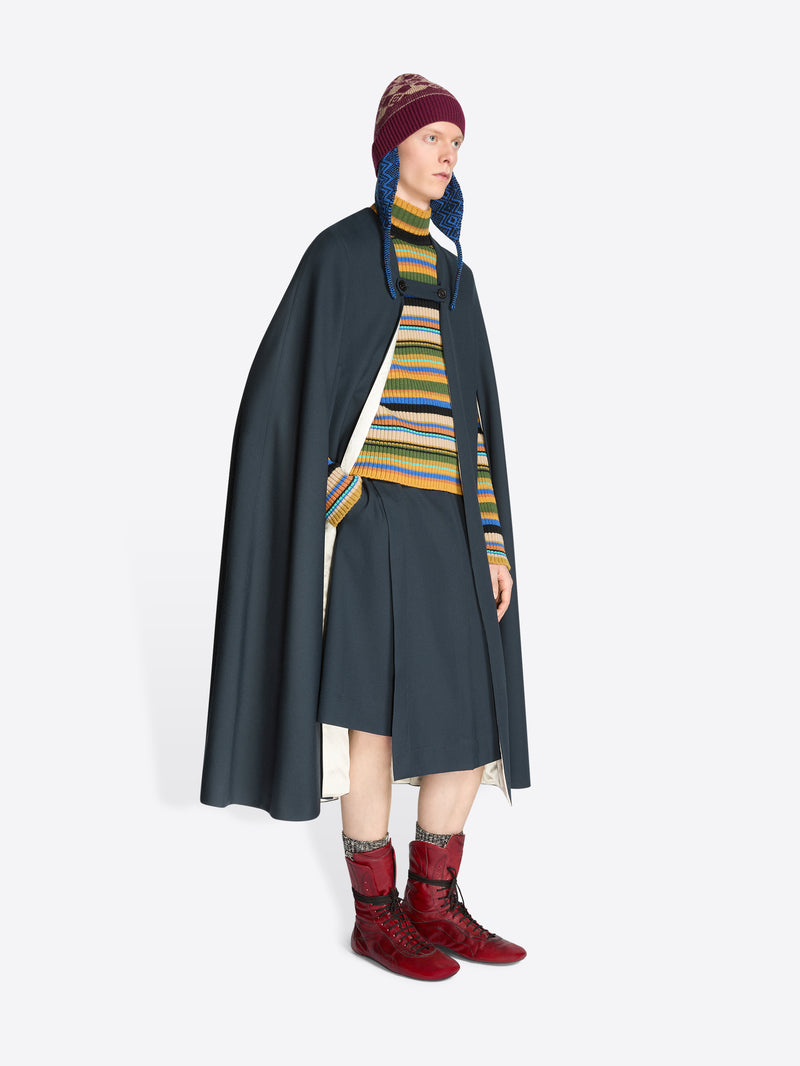 Person wearing a dark cape, striped sweater, skirt, hat and red boots on a white background